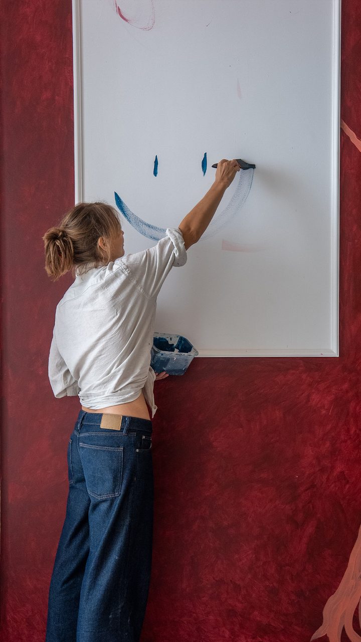 Artist painting a blue crescent onto a white panel set into a deep red wall